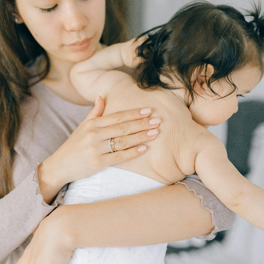 A woman gently holds a baby wrapped in a white towel, softly rubbing the baby's back. The baby's face is turned away, and the woman is looking down with a calm, caring expression.