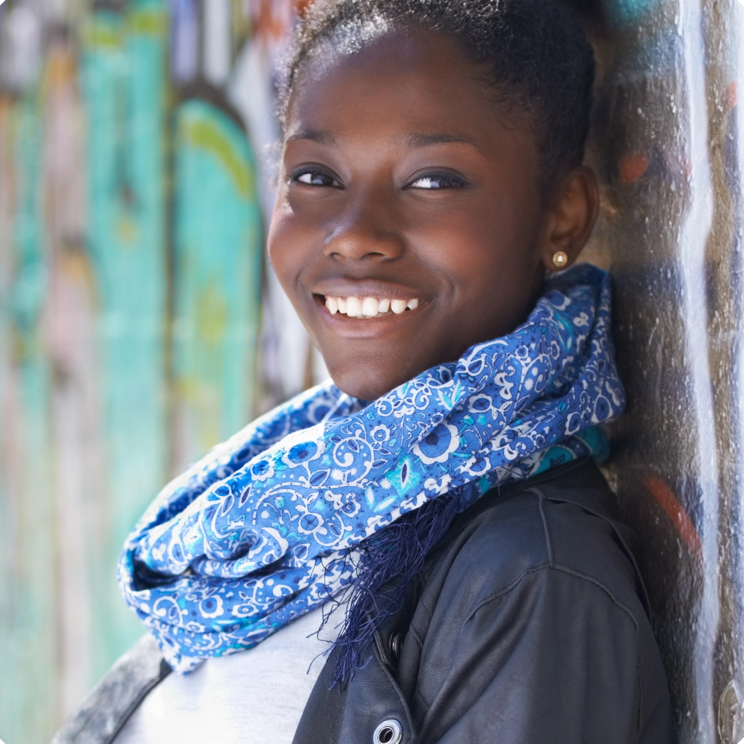 A young woman with dark skin and a bright smile stands against a graffiti-covered wall, wearing a blue patterned scarf and a black jacket.