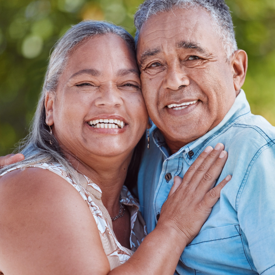 A smiling older couple stands close together outdoors, embracing and looking at the camera. They appear happy and relaxed, with greenery softly blurred in the background.