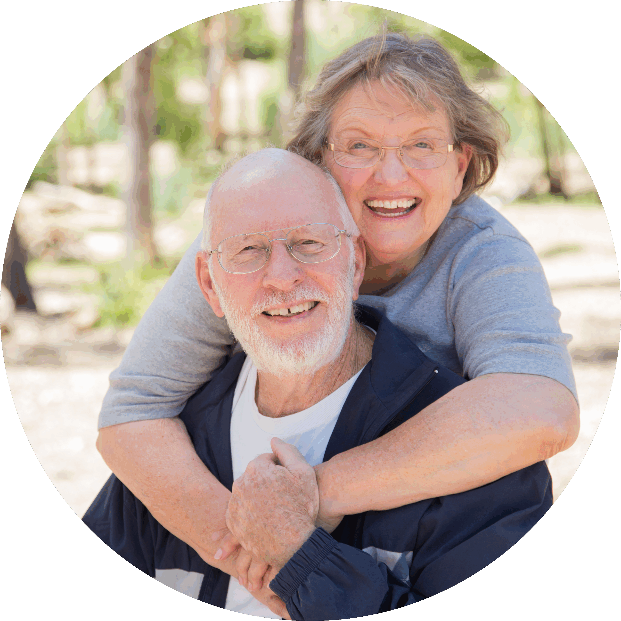 A smiling elderly woman hugs an elderly man from behind. Both are wearing glasses and casual clothes, and they appear happy outdoors on a sunny day.