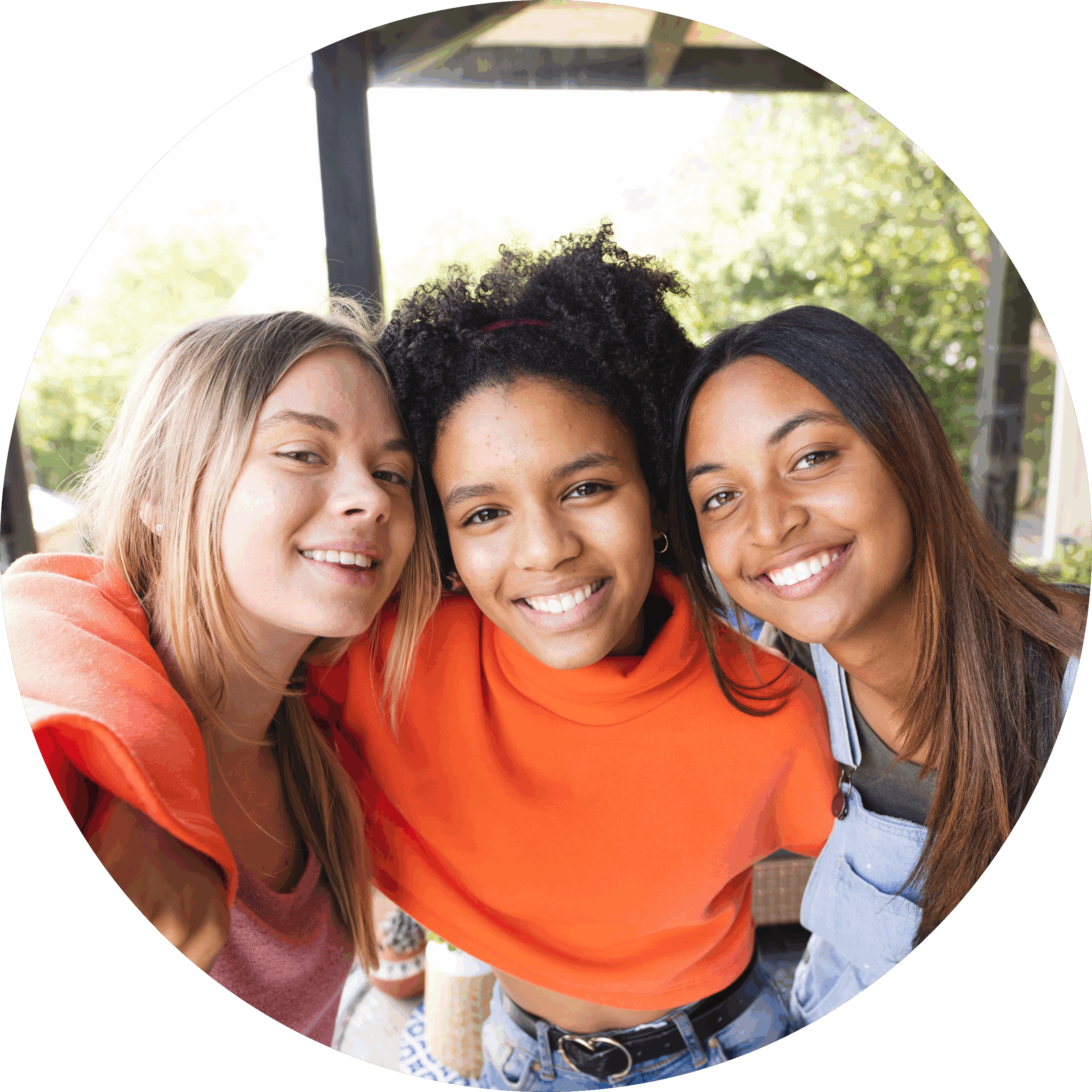 Three young women smiling and posing closely together for a selfie outdoors, with greenery and a wooden structure in the background.