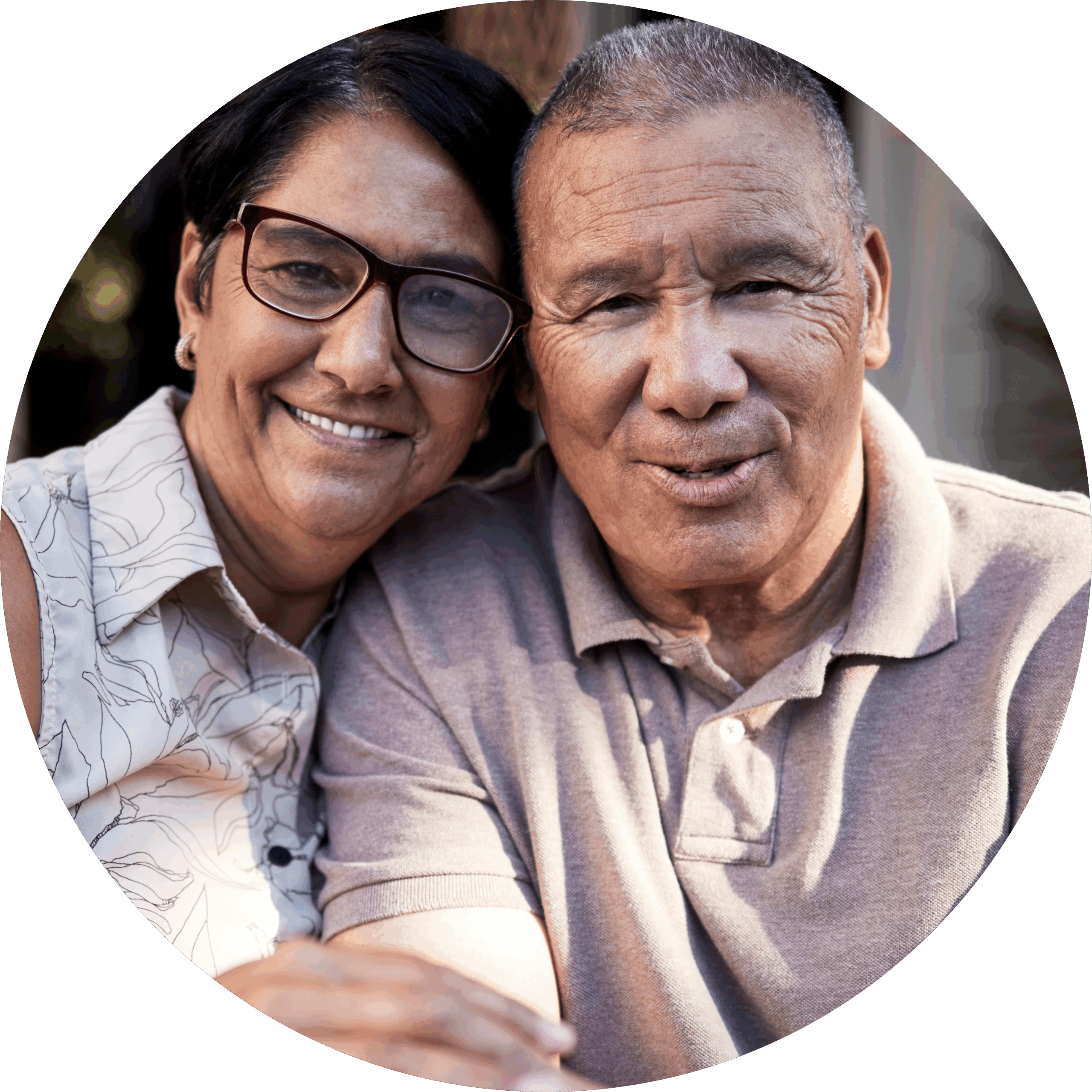 An older couple sits closely together, smiling warmly at the camera. The woman wears glasses and a sleeveless patterned blouse, while the man wears a light polo shirt. They appear happy and content.
